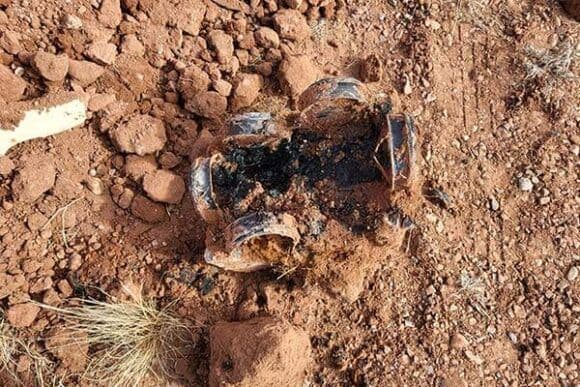 Unidentified burned object partially buried in red dirt with scattered rocks and grass.
