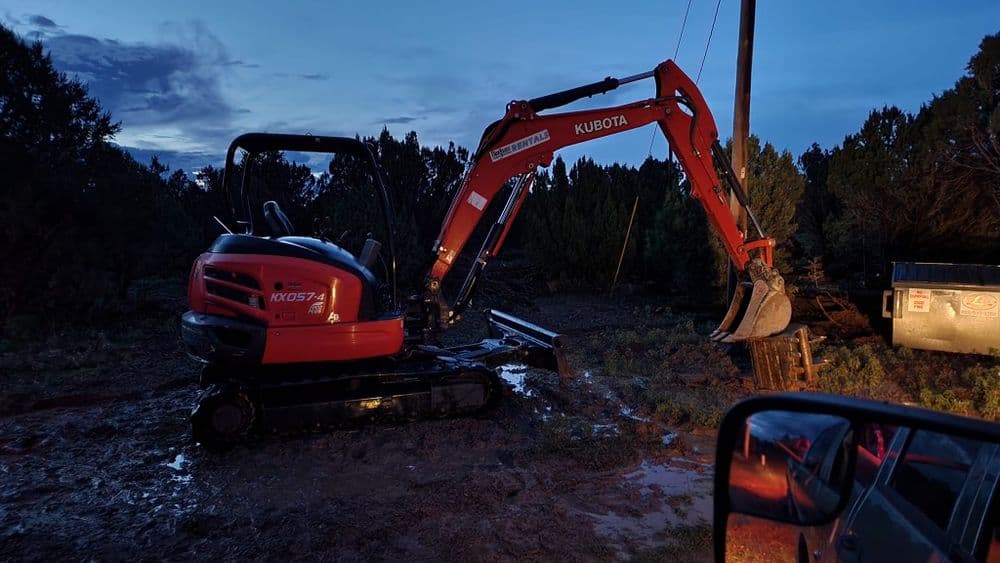 Kubota excavator at dusk, working in muddy terrain surrounded by trees.