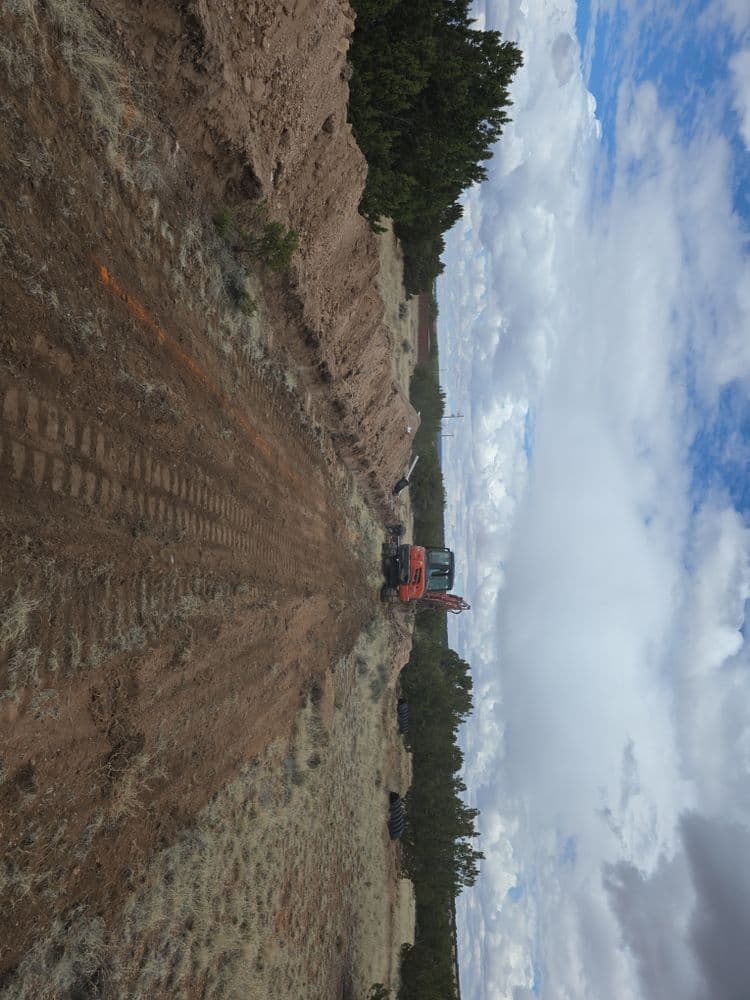 Excavator working on a dirt road in a rural landscape with blue skies and scattered clouds.