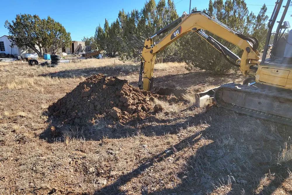 Excavator digging a dirt mound in an outdoor construction site with trees in the background.