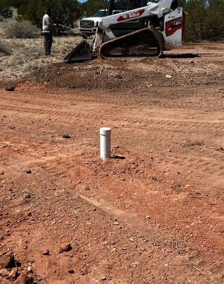 Worker operating a skid steer on a dirt road with a white pipe marker visible.