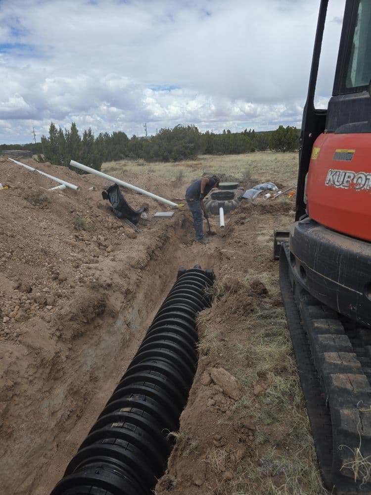 Worker installing drainage pipes in a trench with construction equipment nearby on a rural site.
