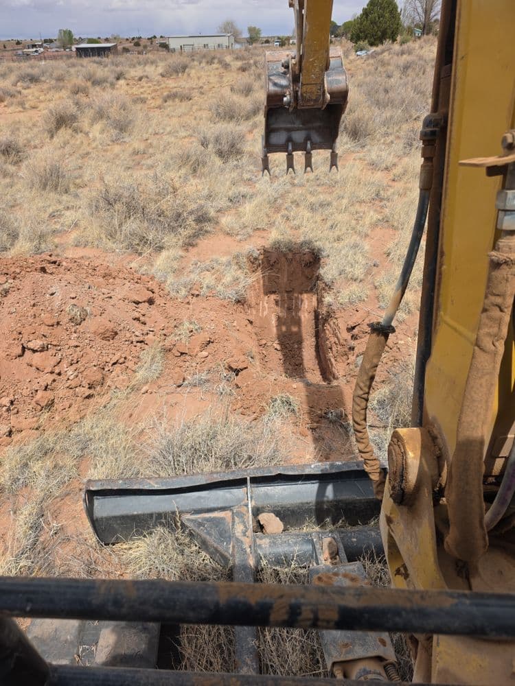 Excavator digging a trench in dry, dusty landscape with sparse vegetation.
