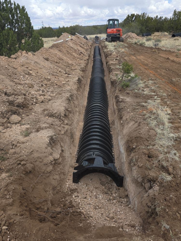 Construction site with a trench, large black drainage pipe, and excavation equipment in the background.