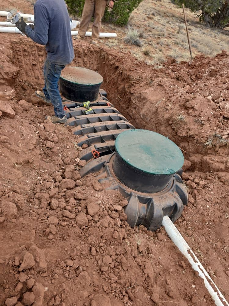Installation of septic tank system in trench with workers and soil. Landscape in background.