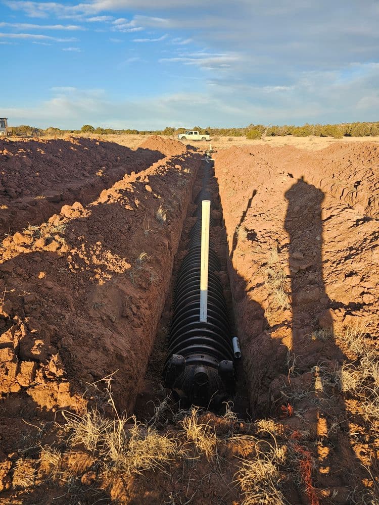 Excavation site with a drainage pipe installed in a trench under blue sky and distant vehicles.