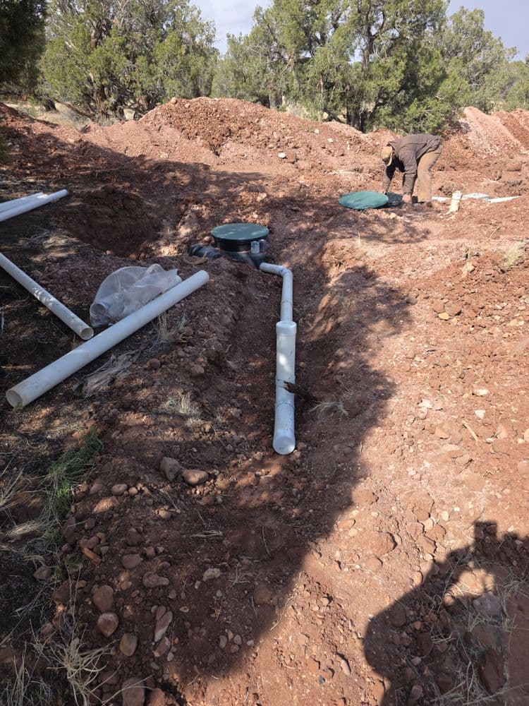 Construction site with PVC pipes in a dirt trench and a worker in the background.