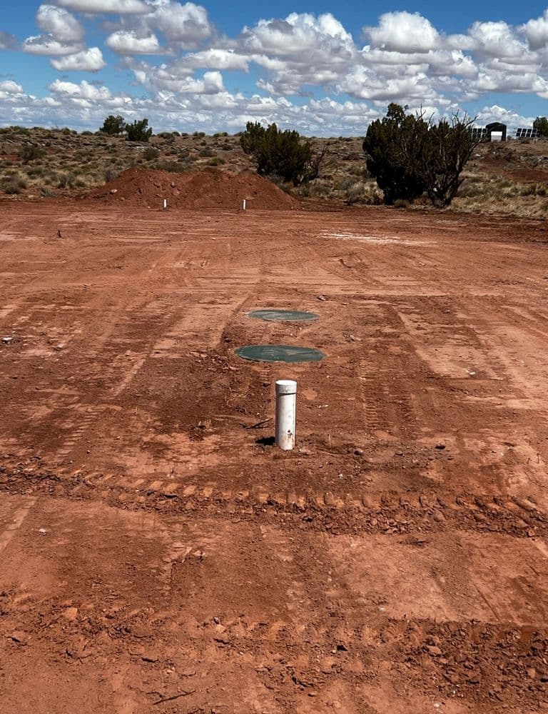 Dry desert landscape with soil, utility covers, and scattered vegetation under a cloudy sky.