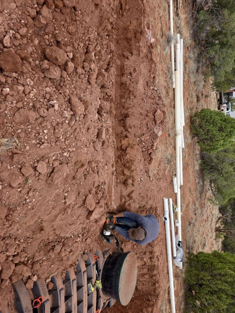 Man working on land excavation with PVC pipes in a red dirt area. Construction site details visible.