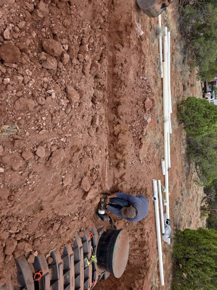Worker installing drainage pipes in a red dirt trench surrounded by construction materials.
