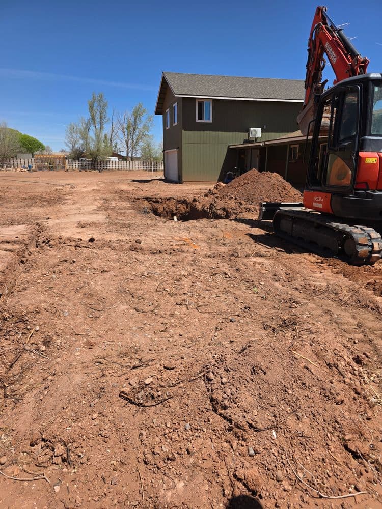 Excavator working on a dirt lot near a residential building under clear blue skies.