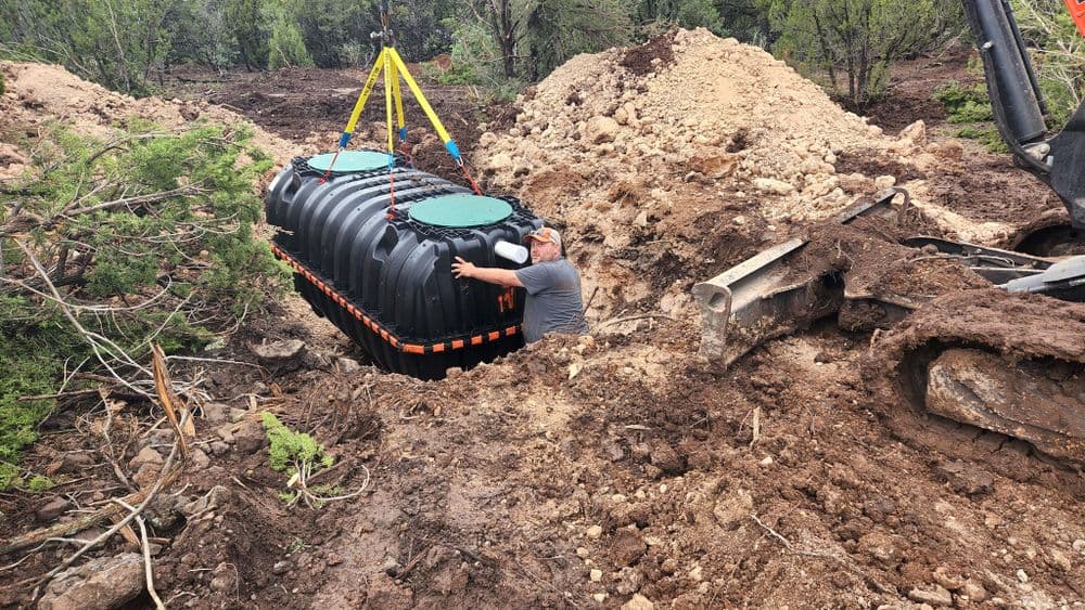 Worker installing a black water storage tank in a trench surrounded by soil and trees.
