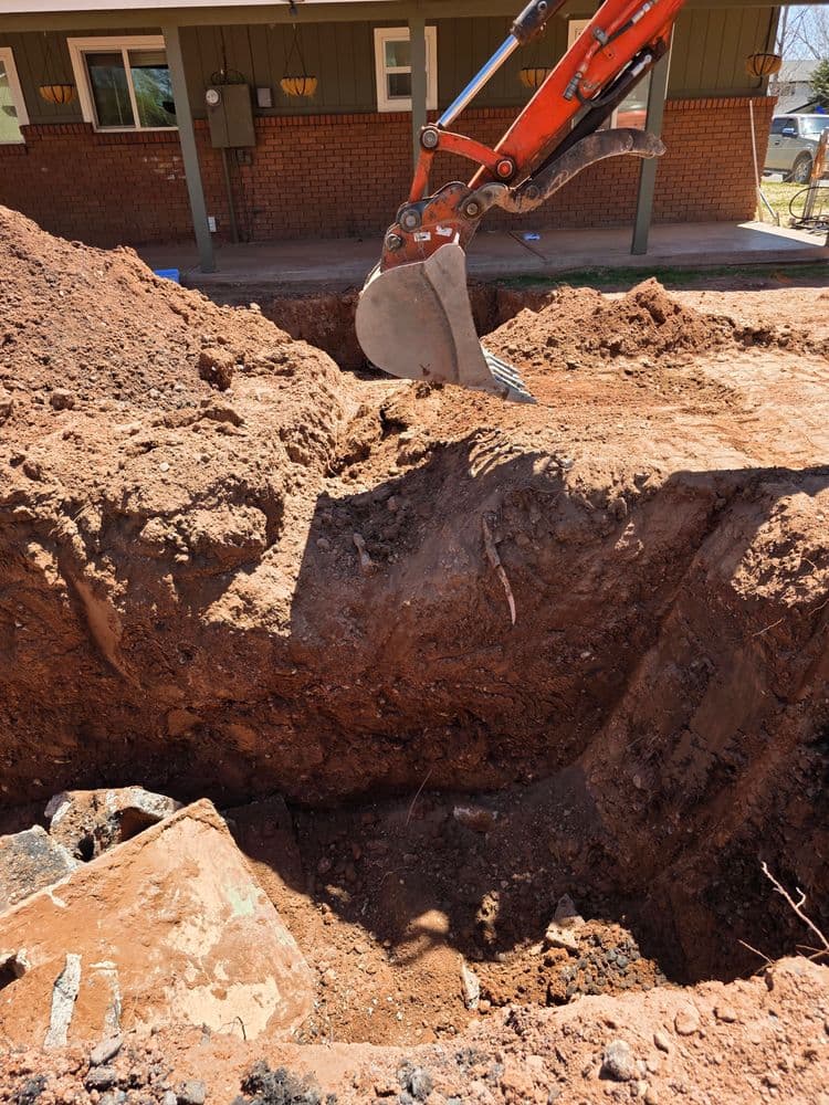 Excavator digging a trench in the ground for construction near a residential building.