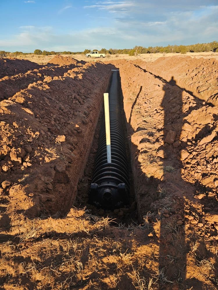 Trench filled with a black drainage pipe, surrounded by earth, under a blue sky.