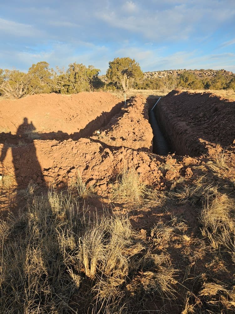 Excavated trench in dirt landscape with shadows and vegetation under blue sky.