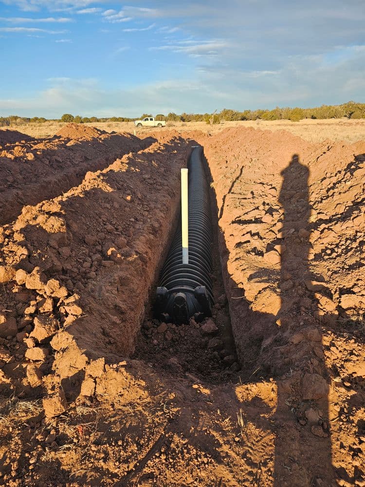 Trench with black piping installed in red soil under a clear sky. Shadow of a person visible.