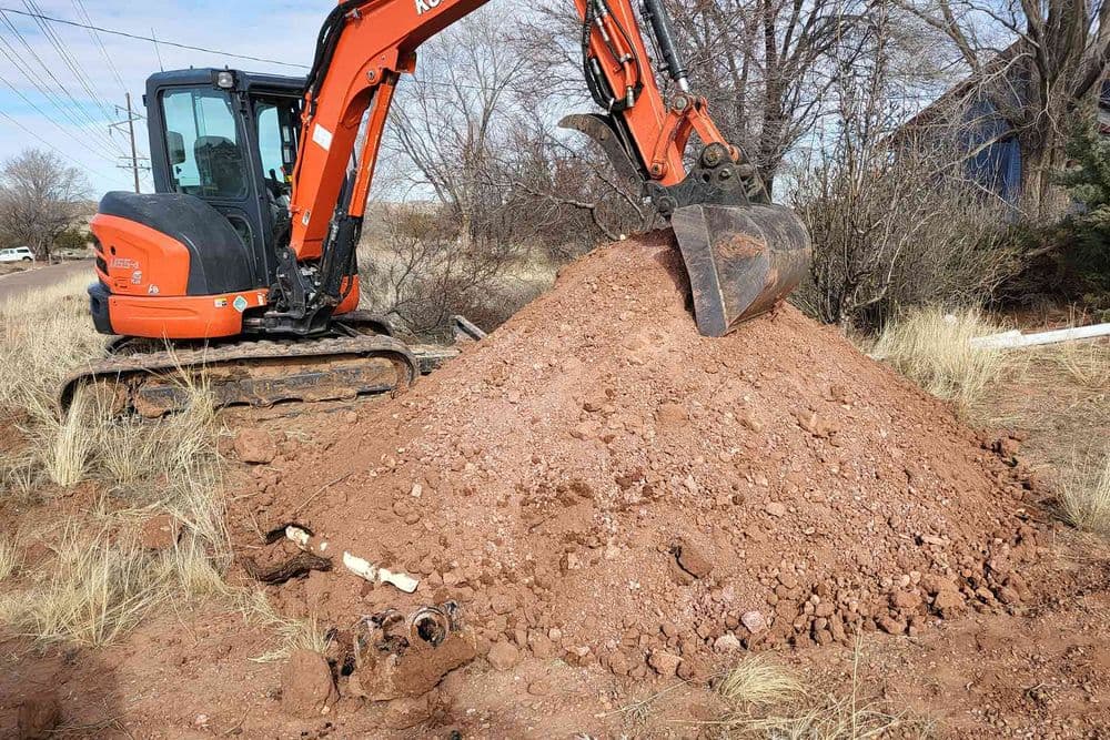 Excavator digging up red soil pile near utility lines in a rural area.