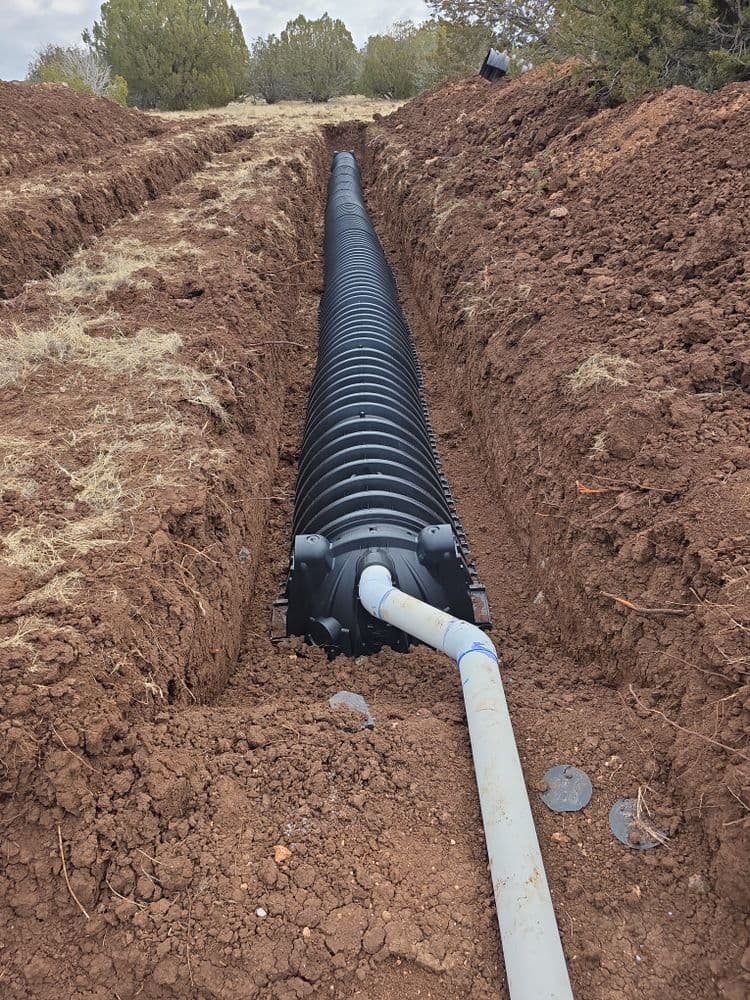 Plastic drainage pipe installed in a trench surrounded by dry earth and sparse vegetation.