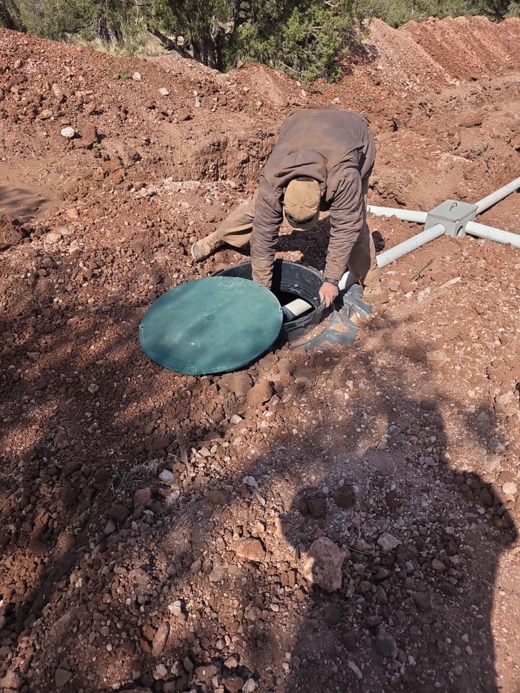 Worker inspecting a green manhole cover in a dirt excavation site.