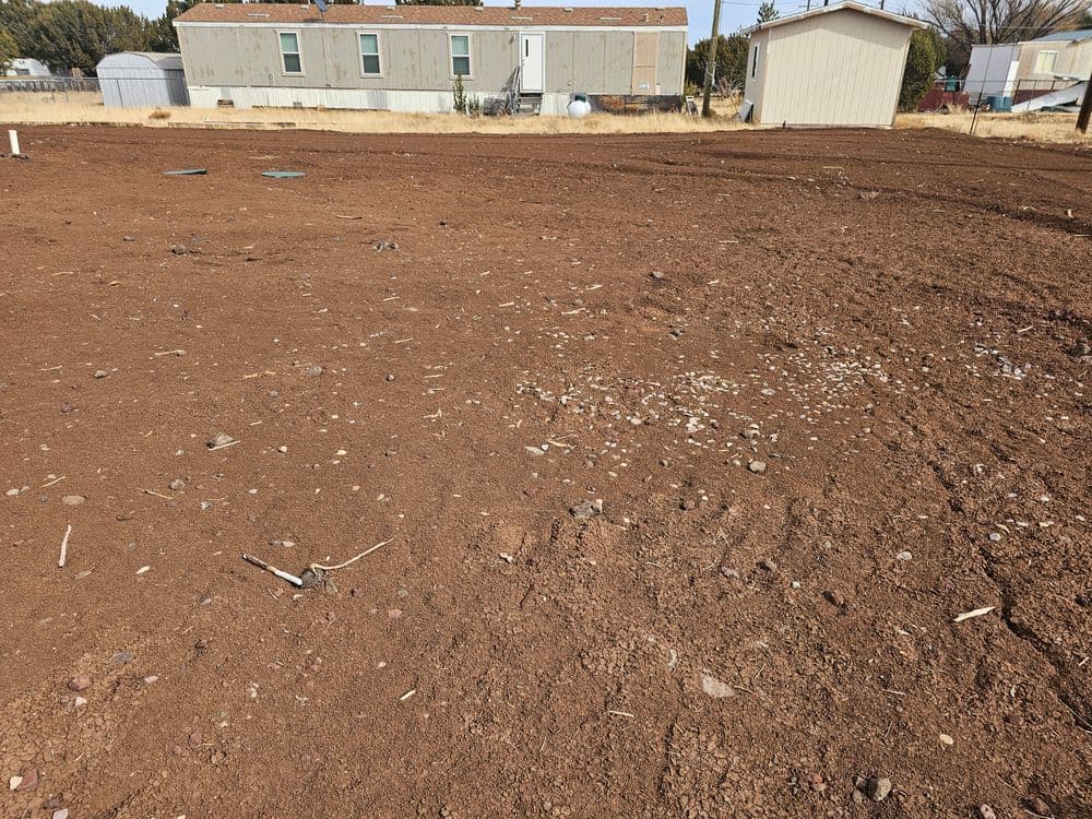 Brown barren land with scattered rocks, mobile homes in the background under clear sky.
