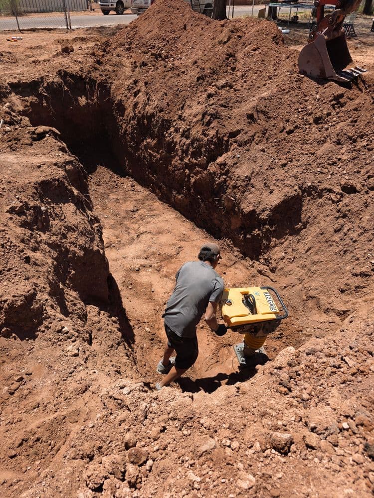 Construction worker operating a compactor in a deep excavation site with brown soil.