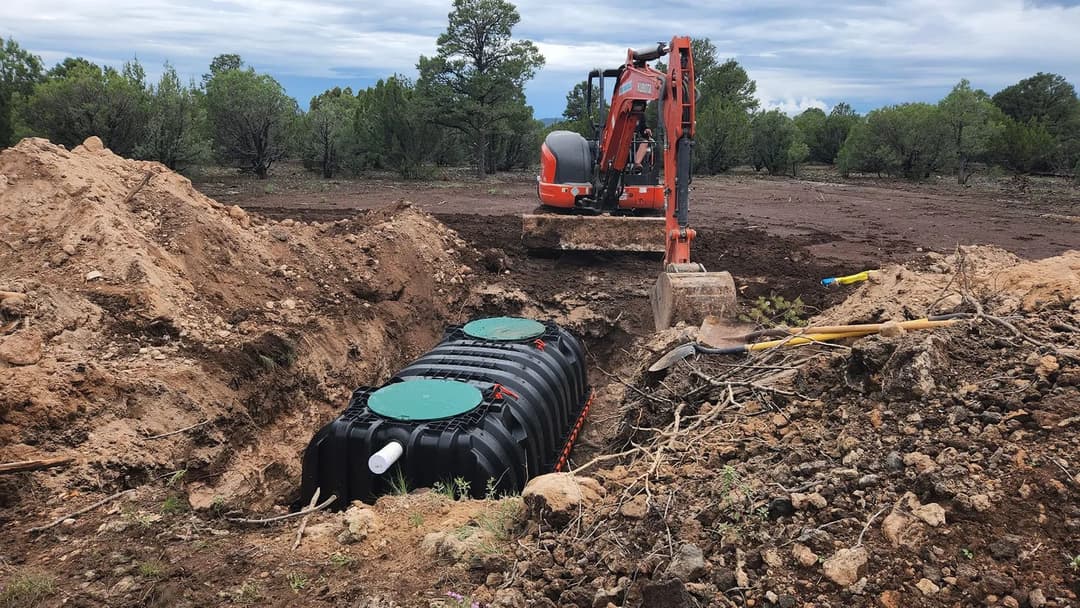 Excavator digging near a buried water tank in a rural landscape with trees.