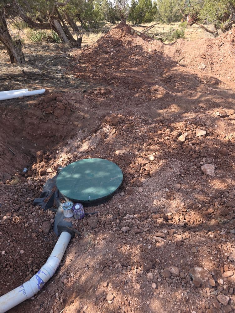 Sewage pump station in a trench with green cover and PVC piping, surrounded by rocky soil.